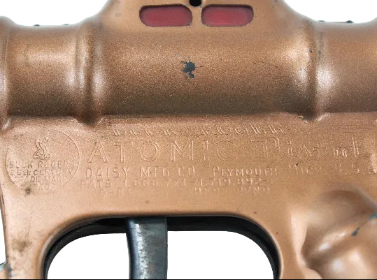 Close-up of a brass toy gun with 'Atomic Model' branding on a light blue background