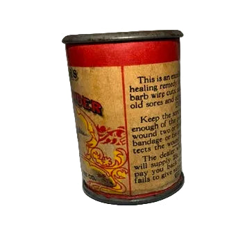 A small cylindrical metal tin with a beige label featuring red and yellow graphics, photographed against a wrinkled white fabric background.