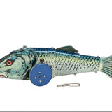 A vintage metal fish-shaped toy with blue wheels, featuring a blue and green painted body, red eye, and a small metal key or accessory placed in front, photographed against a white fabric background.