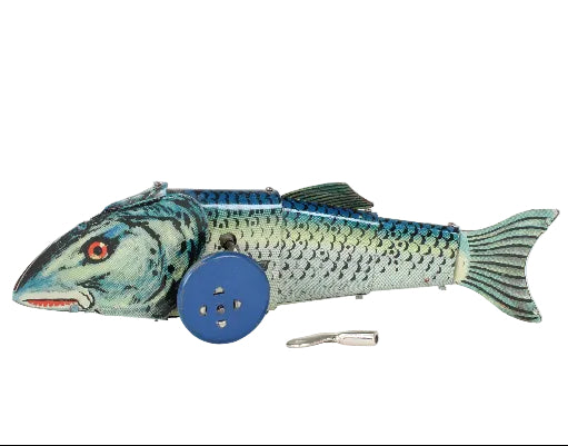 A vintage metal fish-shaped toy with blue wheels, featuring a blue and green painted body, red eye, and a small metal key or accessory placed in front, photographed against a white fabric background.