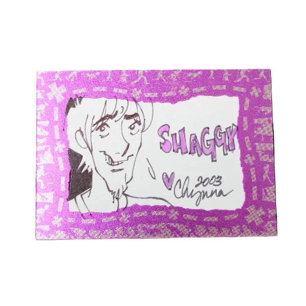A rectangular printed card or sticker with a pink and purple patterned border, featuring a black line drawing of a smiling anime-style character and the word 'SHAGGY' with a heart and the name 'Chiguma' below, placed on a dark textured background.