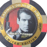 A circular 007 Casino L'Or Noir limited edition betting chip featuring a portrait of Pierce Brosnan with the text 'The World Is Not Enough 007' on a dark background.
